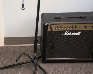 A black guitar stand and a Marshall amplifier are placed on a gray carpet next to a light blue wall.