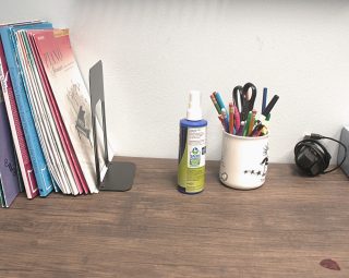 A desk with books, a spray bottle, cup of pens and scissors, headphones, and a tissue box against a light wall.