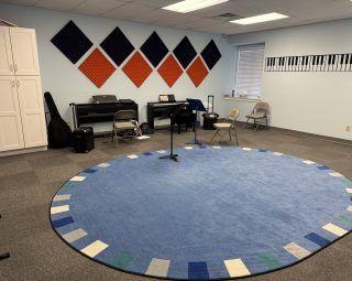 Music classroom with pianos, chairs, and a blue rug; wall has sound panels and piano key decal.