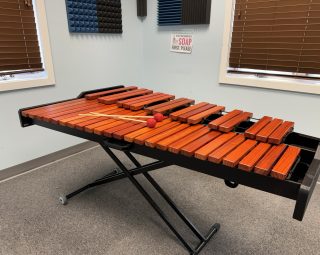 A marimba with two mallets rests on a black stand in a music practice room with brown blinds.