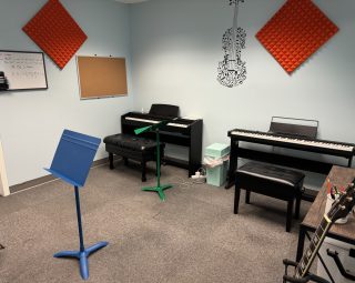 Music classroom with two keyboards, music stands, wall art, acoustic panels, and a guitar on a stand in the corner.