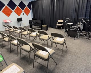 Rows of chairs face a keyboard, drum set, and music stands in a room with soundproofing panels and black curtains.