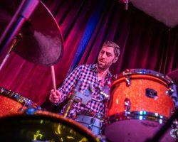 A man in a plaid shirt plays an orange drum set on stage with dark red curtains in the background.
