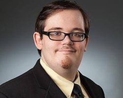 Man wearing glasses, a suit jacket, yellow shirt, and black tie, facing forward with a neutral expression.