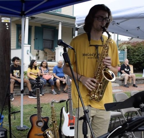 A person in a yellow shirt plays saxophone under a canopy. Several people sit and watch on benches in the background.