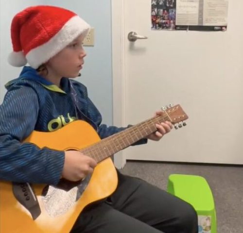 Boy wearing a Santa hat plays an acoustic guitar while seated in a room with a music poster on the door.
