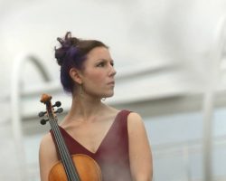 Woman in a sleeveless dress holds a violin outdoors, standing near water with a bridge in the background.