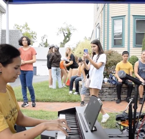Person plays keyboard outdoors while people watch, some sitting and some standing, on a residential street.