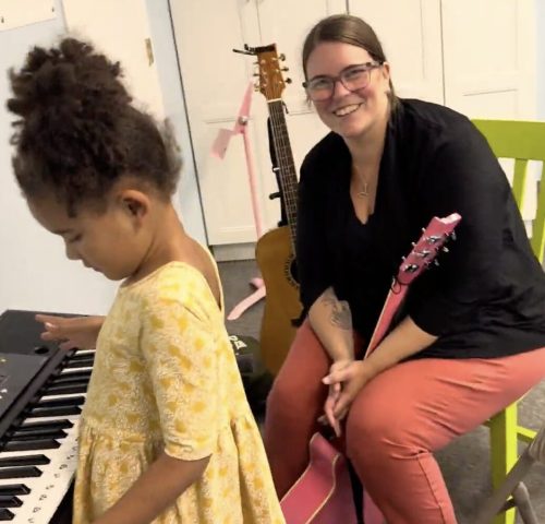 A woman with a pink guitar smiles at a girl playing a keyboard in a music room.