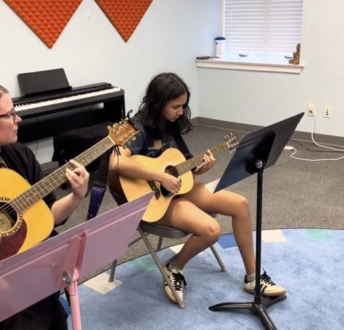 Two people sit in a music room playing acoustic guitars, with sheet music stands and a keyboard in the background.