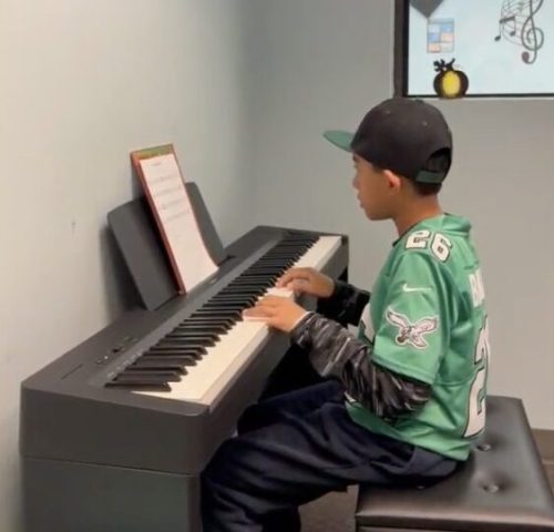 A boy in a sports jersey and cap plays an electric keyboard while reading sheet music in a small music room.