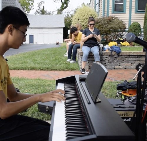 Young man plays electric piano outdoors while a small group of people sit nearby, some looking at their phones.