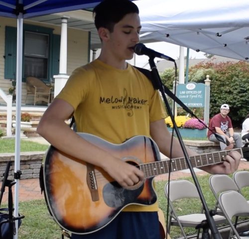 Teen wearing a yellow shirt plays acoustic guitar and sings into a microphone at an outdoor event under a canopy.