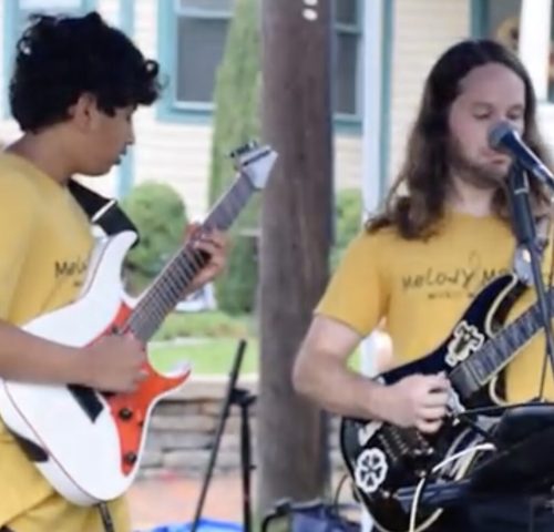 Two musicians wearing yellow shirts play electric guitars and sing outdoors, with houses and greenery in the background.
